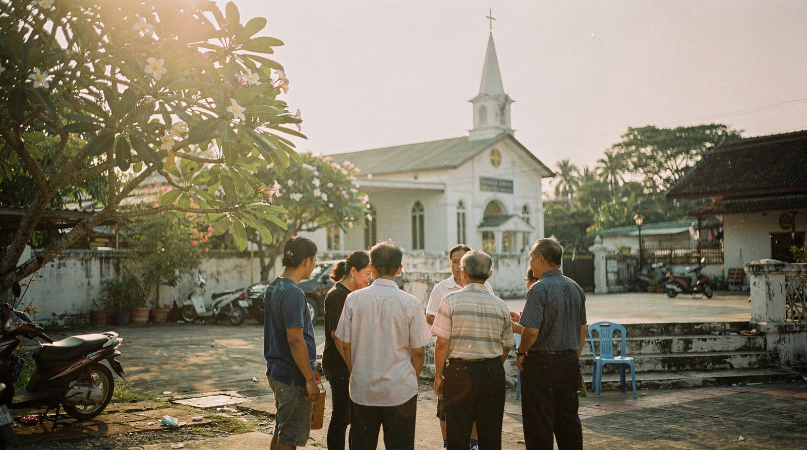 Filipino parishioners talking in a Bangkok church courtyard at golden hour, seen from behind.