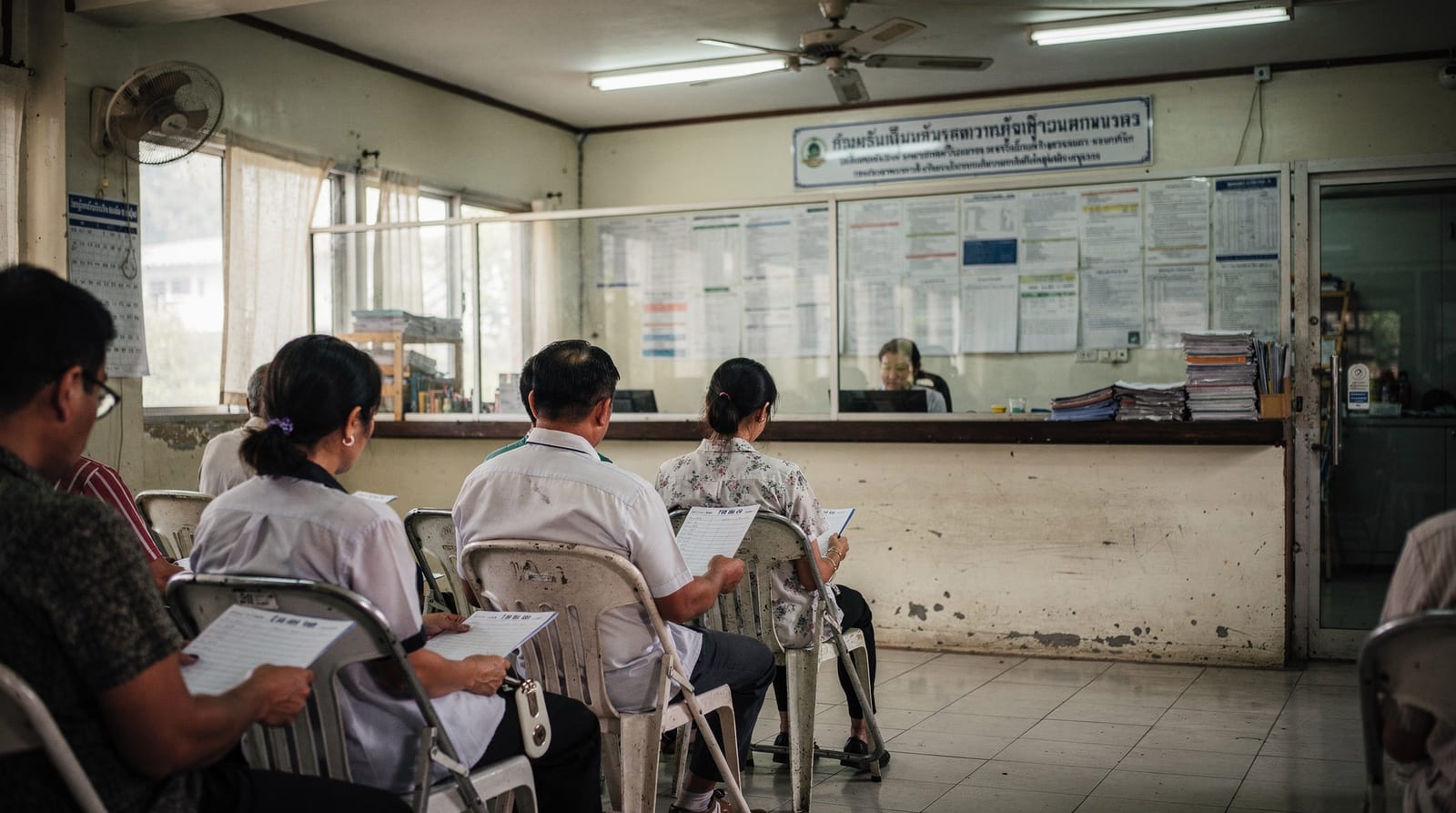 People wait on plastic chairs holding document folders and queue tickets inside a Thai Department of Employment office, seen from behind, facing a glass-partition counter.