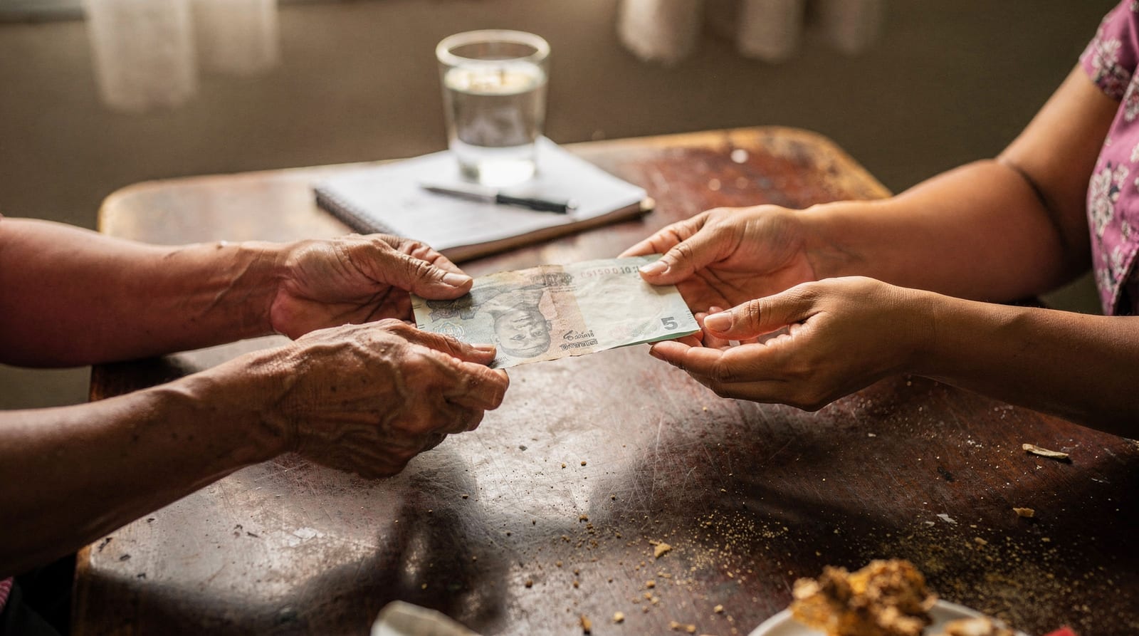 Two pairs of hands exchanging a baht banknote across a table, showing a monthly wage being paid by hand.