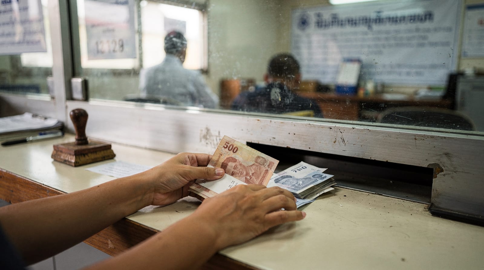 A pair of hands counts Thai baht banknotes at a cashier window with a glass partition and a rubber stamp.