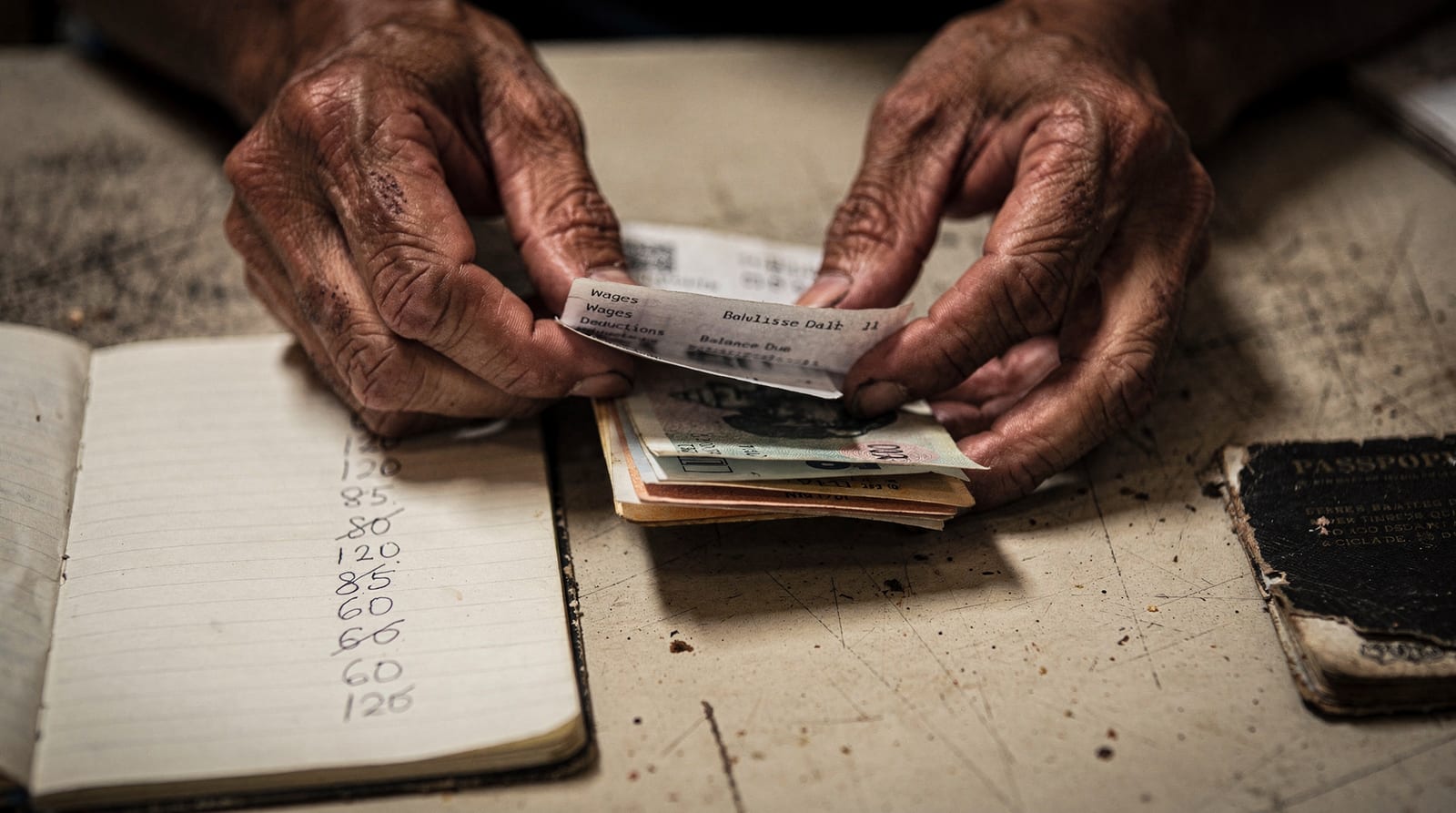 A migrant worker's weathered hands hold a payslip and a thin stack of banknotes beside a notebook of handwritten deductions, no face visible.